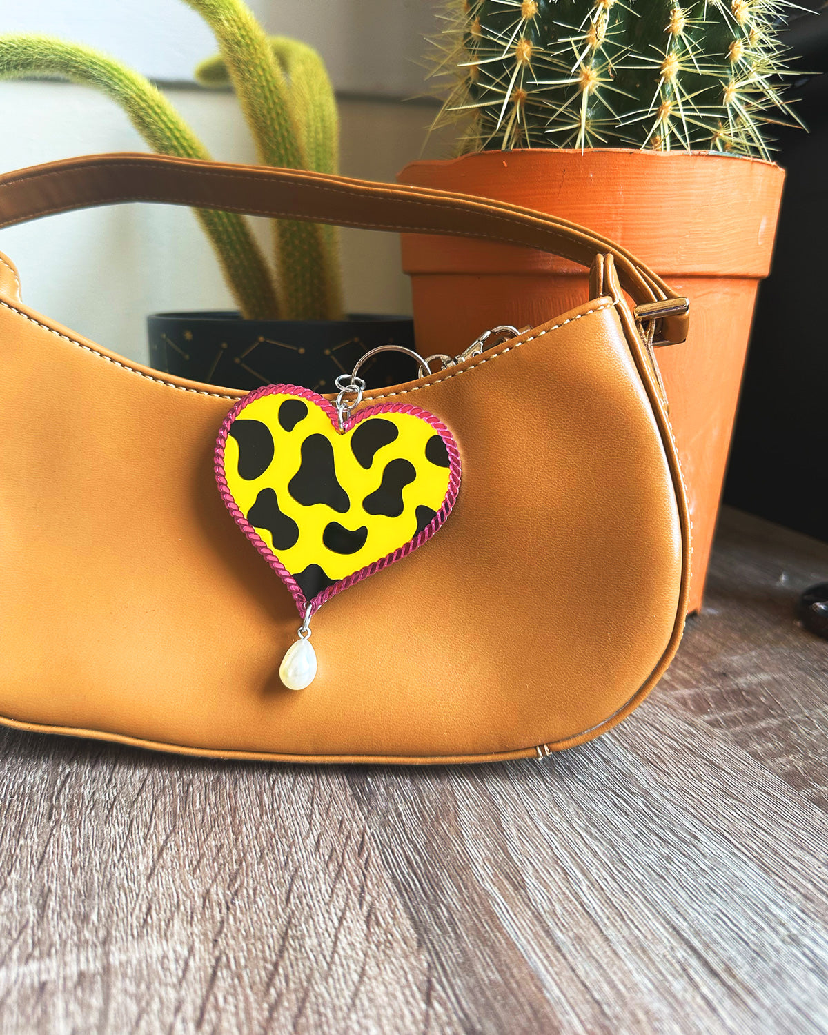 Brown handbag with a heart-shaped bag charm keychain featuring a yellow background and black patterns on a wooden surface.