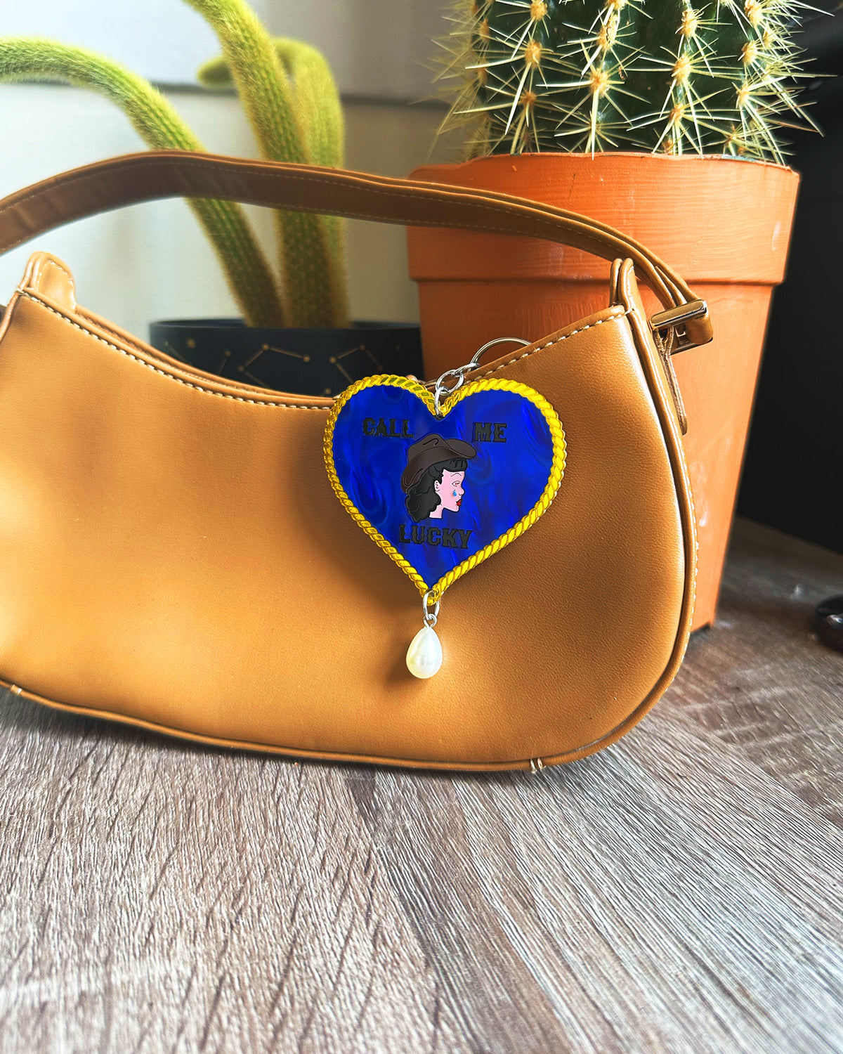 Brown handbag with a blue heart-shaped bag charm keychain featuring a cartoon character on a wooden surface.