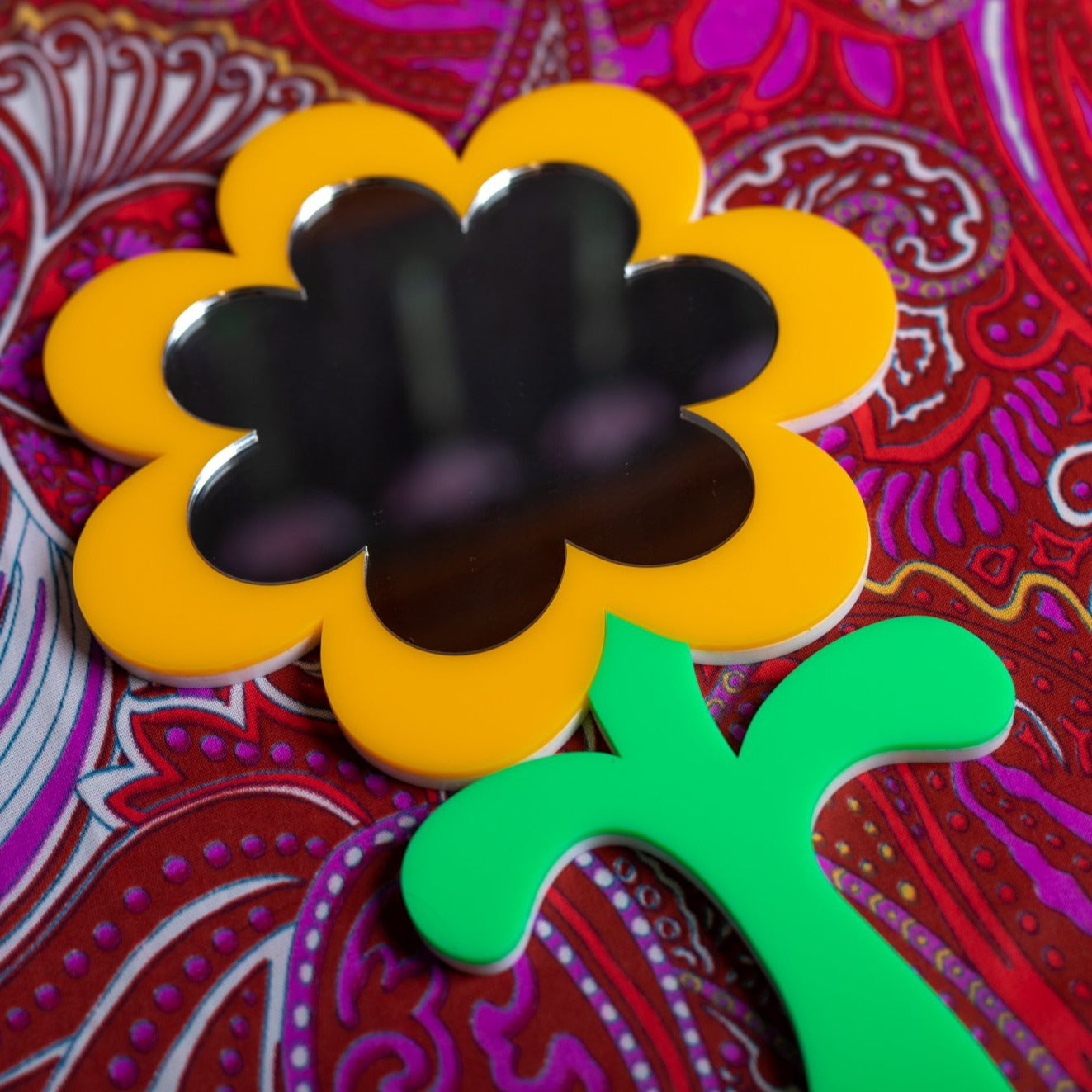 Close up of the new MindFlowers handheld mirror. The handle is a bright green color, shaped like a flower stem. The main mirror is shaped as a daisy with an outer shape featuring a bright canary color.